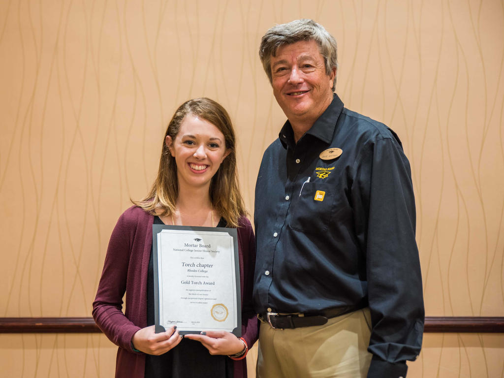 a young white woman smiling and proudly holding an award that she received from an older white male that is also pictured