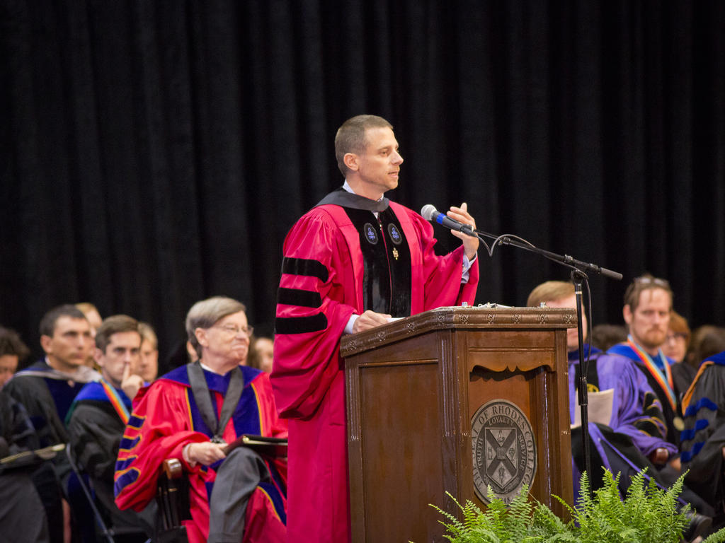 a professor in academic robes giving a speech at a wooden podium