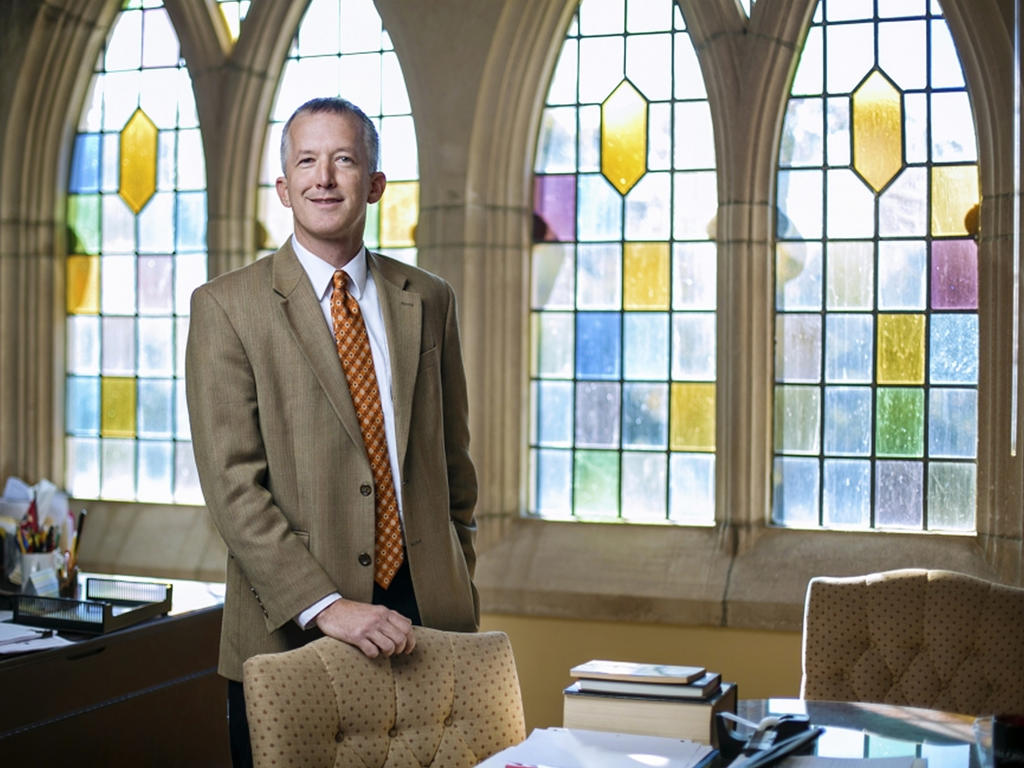 a middle aged while male professor standing near a desk in a room with stained glass windows