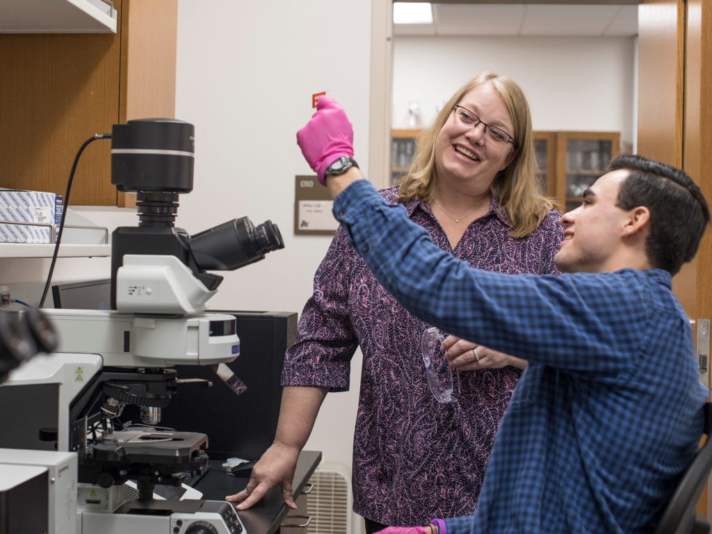A female professor stand over a male student holding up a slide in front of a microscope