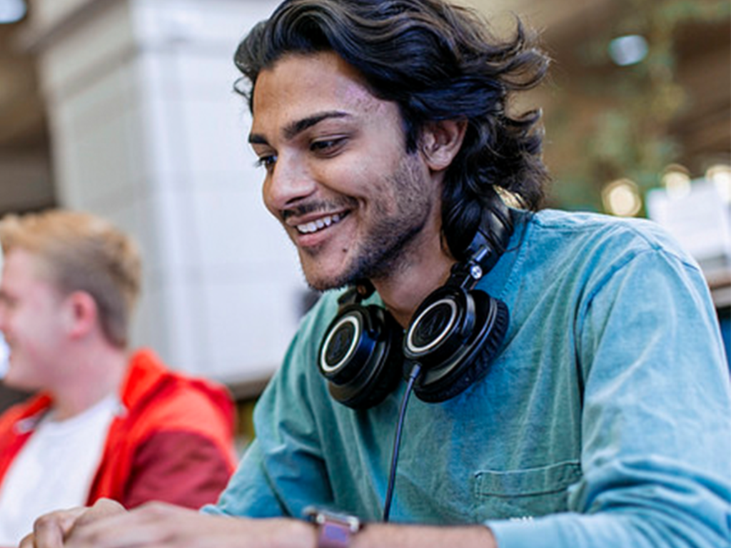 a young Indian male student with headphones around his neck, working and smiling