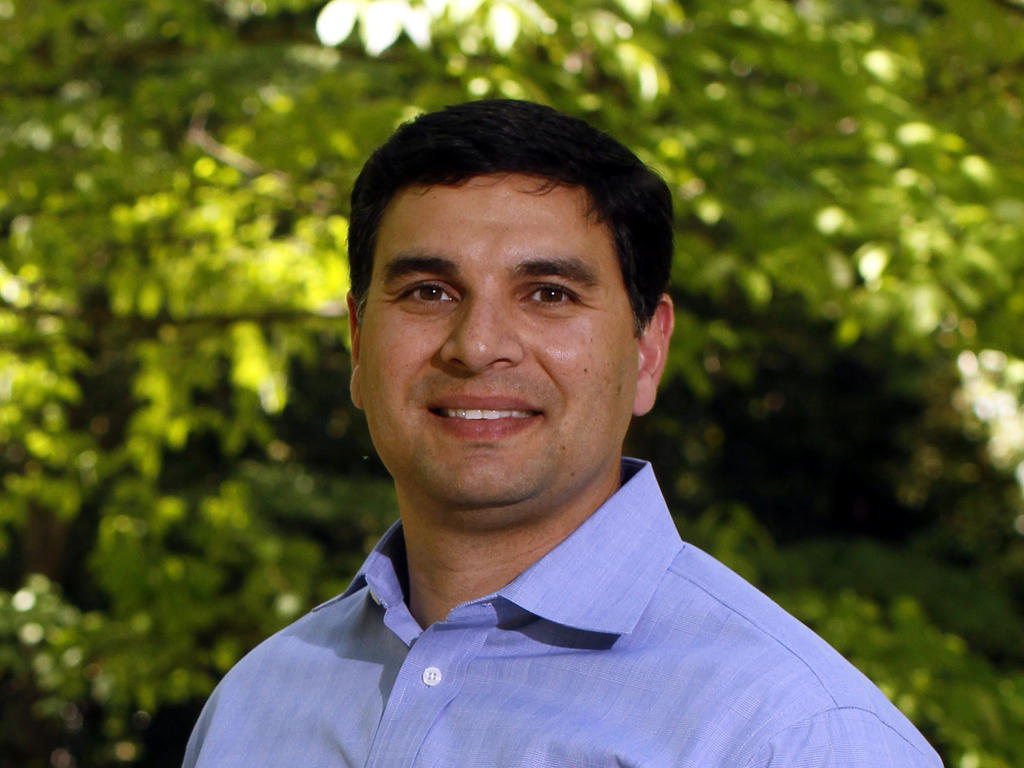 Close shot of a tan, middle-aged, male professor standing in front of greenery