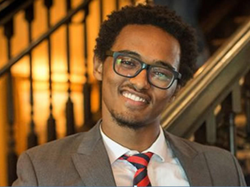 A closeup of an African American male student wearing rectangular glasses, a big smile, and a red and black striped tie