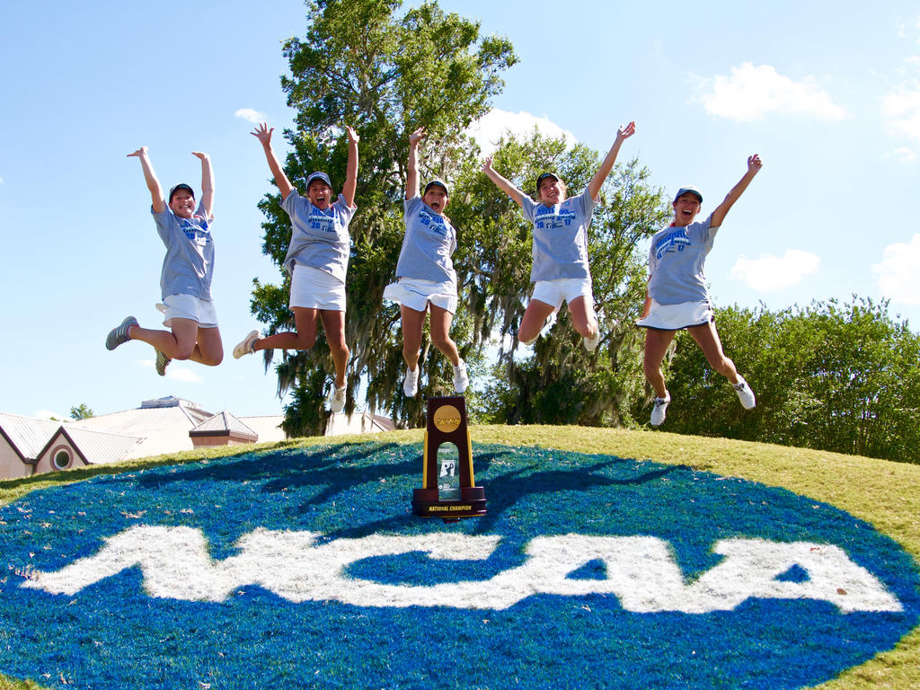 five young women dressed in golfing attire jump victoriously celebrating the trophy at the front of the image