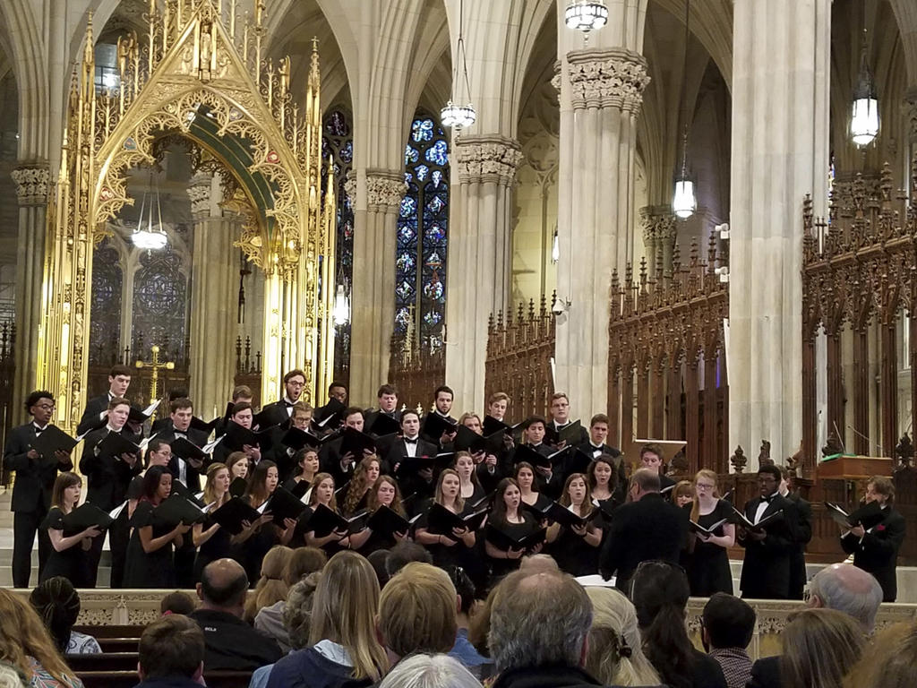 A group of college-aged males and females standing on the altar of a cathedral facing their conductor